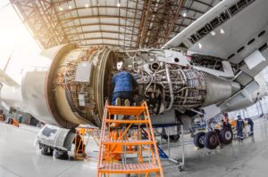 A mechanic sits on an orange ladder as he repairs the engine of a white passenger aircraft